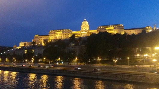 kongeslottet i budapest set fra kædebroen Széchenyi som lyser op i mørket. Slottet ligger bagerst men dens skær ligger i det flotte vand foran.